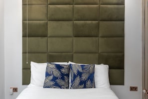 Una cama con cabecera capitonada, ropa blanca y dos almohadones con estampado de hojas.