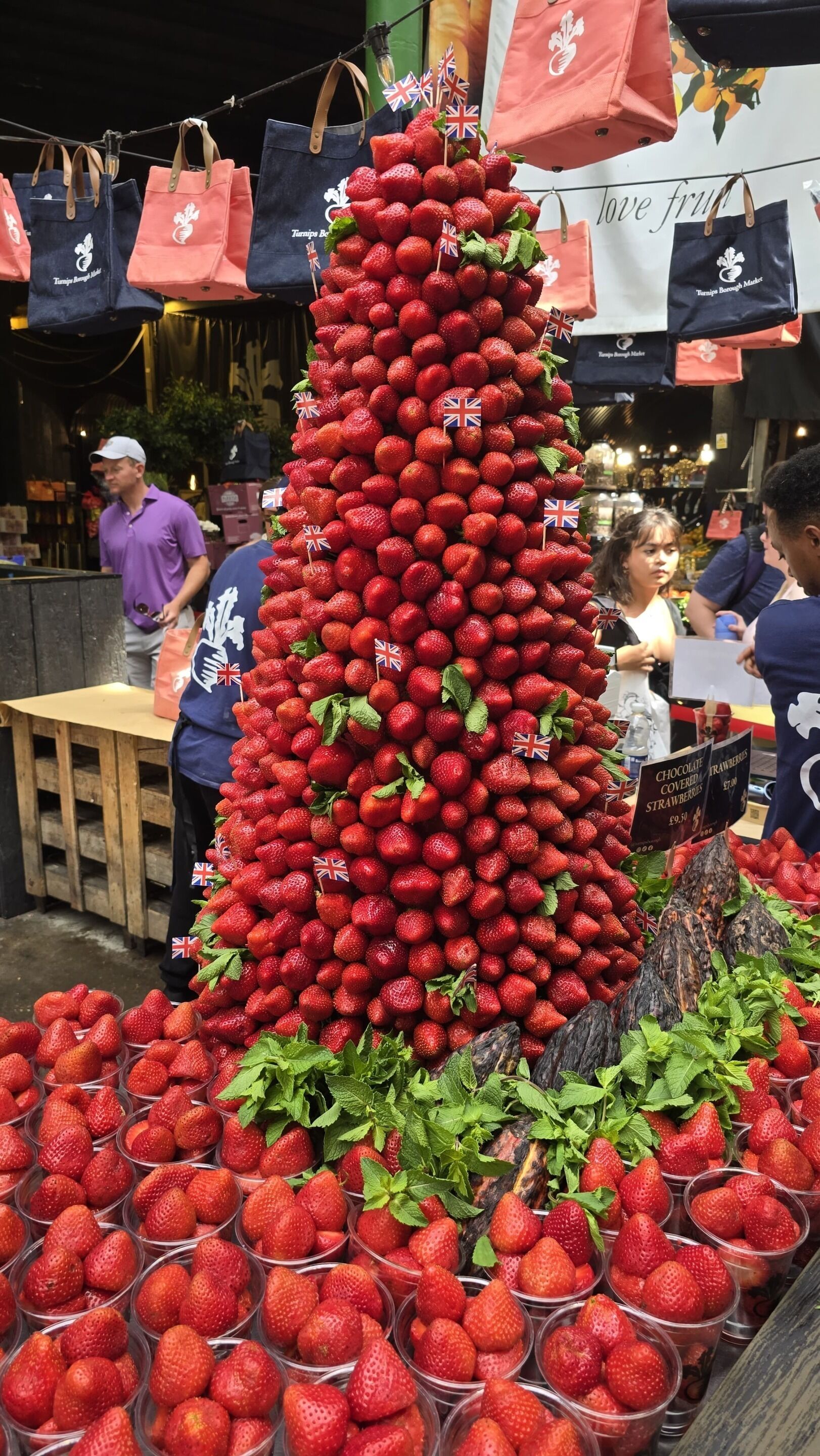 A display of strawberries arranged in a pyramid shape with small flags, surrounded by baskets and a sign indicating a price of £9.50.