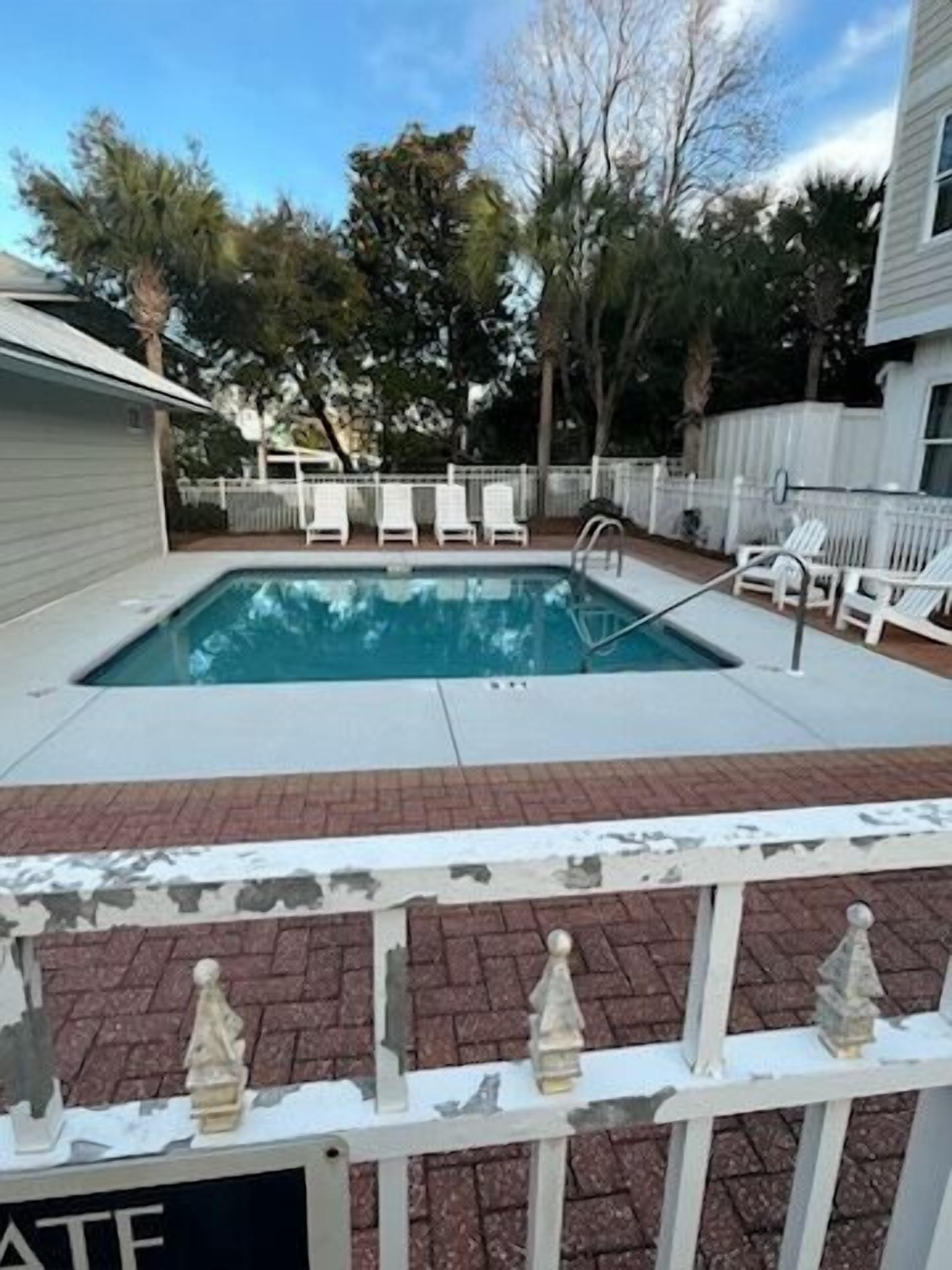 A rectangular swimming pool with a white deck, surrounded by a white fence and trees.