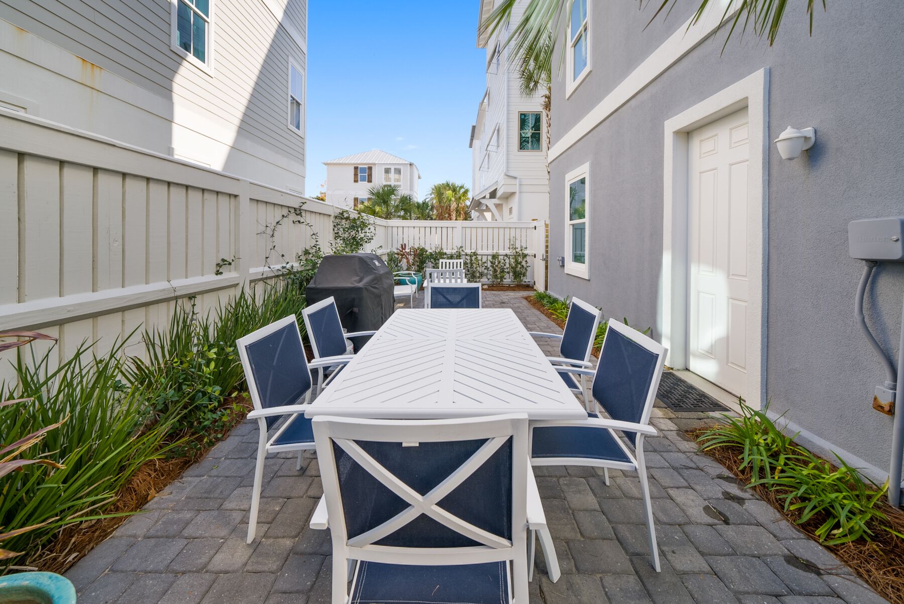 A patio with a white table and blue chairs, surrounded by plants and a white fence.