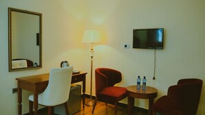 Camera d'albergo con scrivania, due sedie, una lampada, televisore e un tavolino con bottiglie d'acqua.
