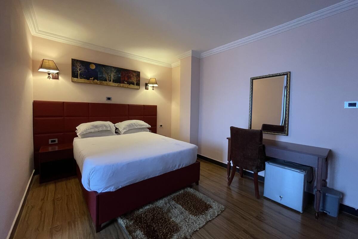 A hotel room with a bed, bedside lamps, a desk with a mirror, and a small refrigerator.