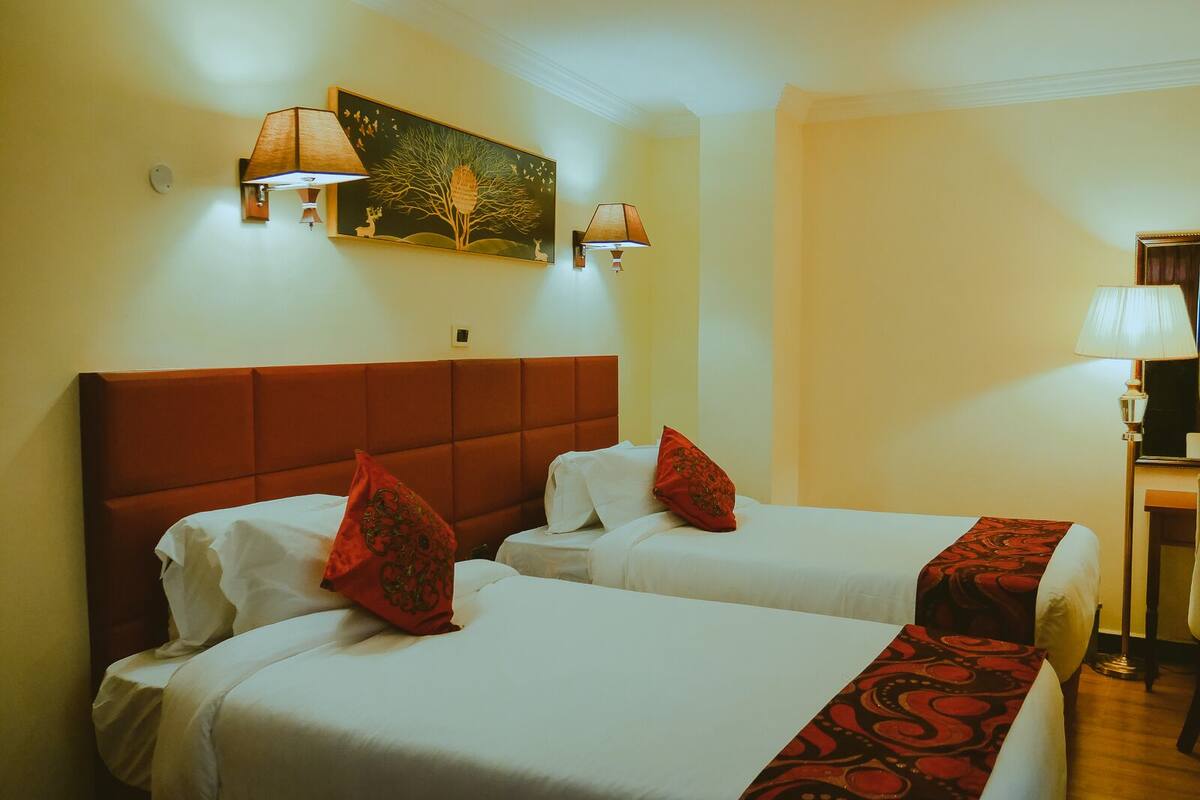 A hotel room with two beds, each with white linens and red decorative pillows. There is a painting on the wall above the beds, and two wall-mounted lamps.