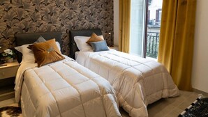 A hotel room with two beds, a patterned wallpaper, a window with yellow curtains, and a view of buildings outside.