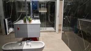 A modern bathroom with a large white sink, a glass-enclosed shower, and a marble-tiled wall.