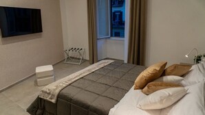 A bedroom with a bed, a TV mounted on the wall, a window with curtains, and a bedside table with a lamp.