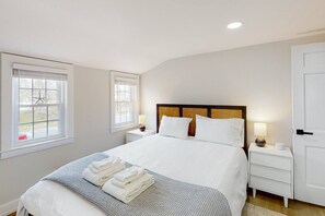 A neatly made bed with white linens and a folded towel, two bedside tables with lamps, and a window with a view of trees. - Modern Cape Home in Hyannis walking distance to the beach & town! (Barnstable)