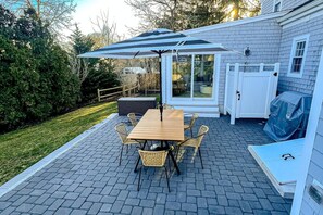 A patio with a wooden table and chairs, a covered umbrella, and a glass door leading to a covered area. - Modern Cape Home in Hyannis walking distance to the beach & town! (Barnstable)