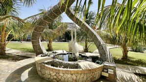 A stone fountain with a white bird sculpture in a circular stone basin, surrounded by palm trees and a grassy area. - Cloud Cloud Farm Resort (Izki)