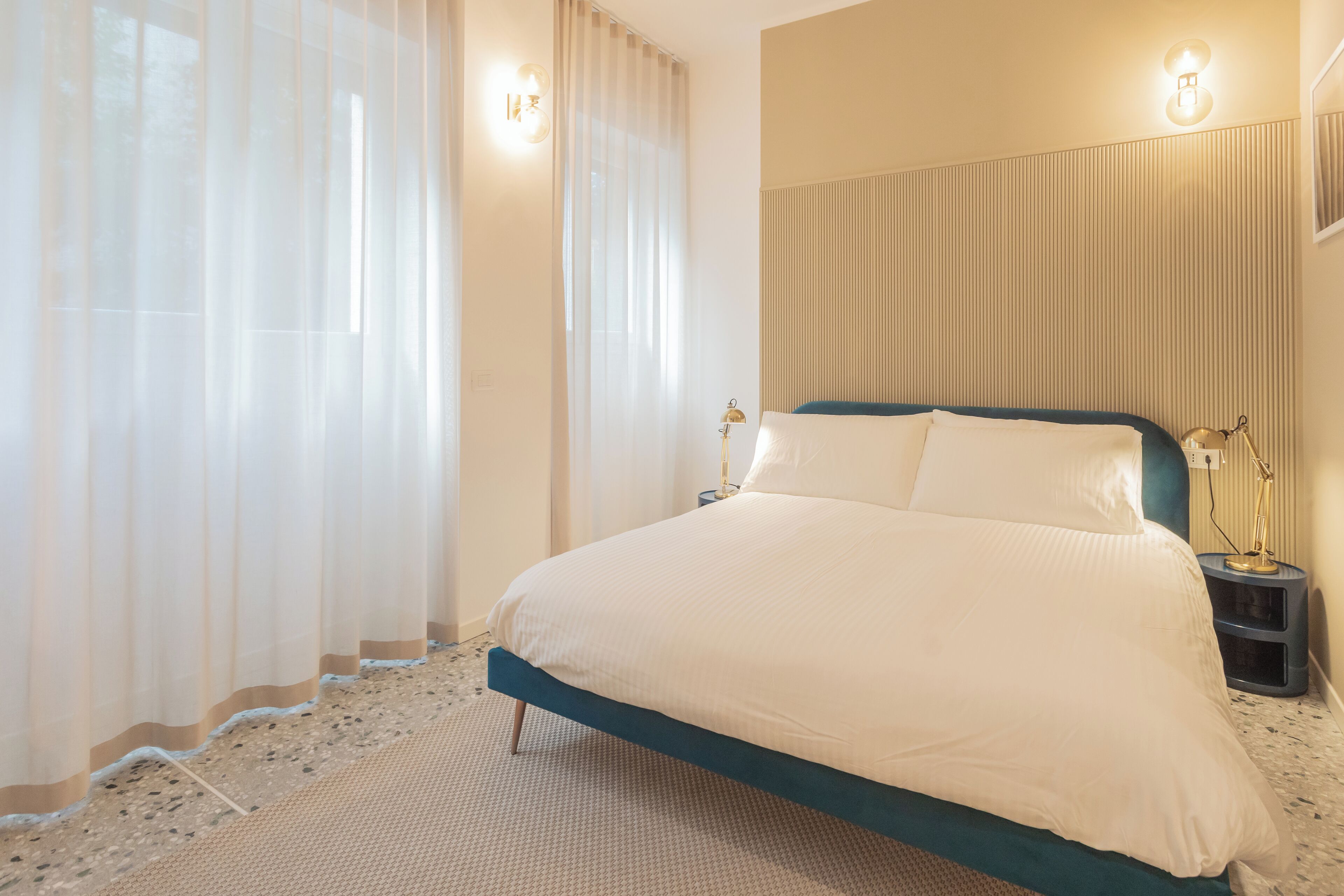 A neatly made bed with a blue headboard, a bedside lamp, and sheer curtains.