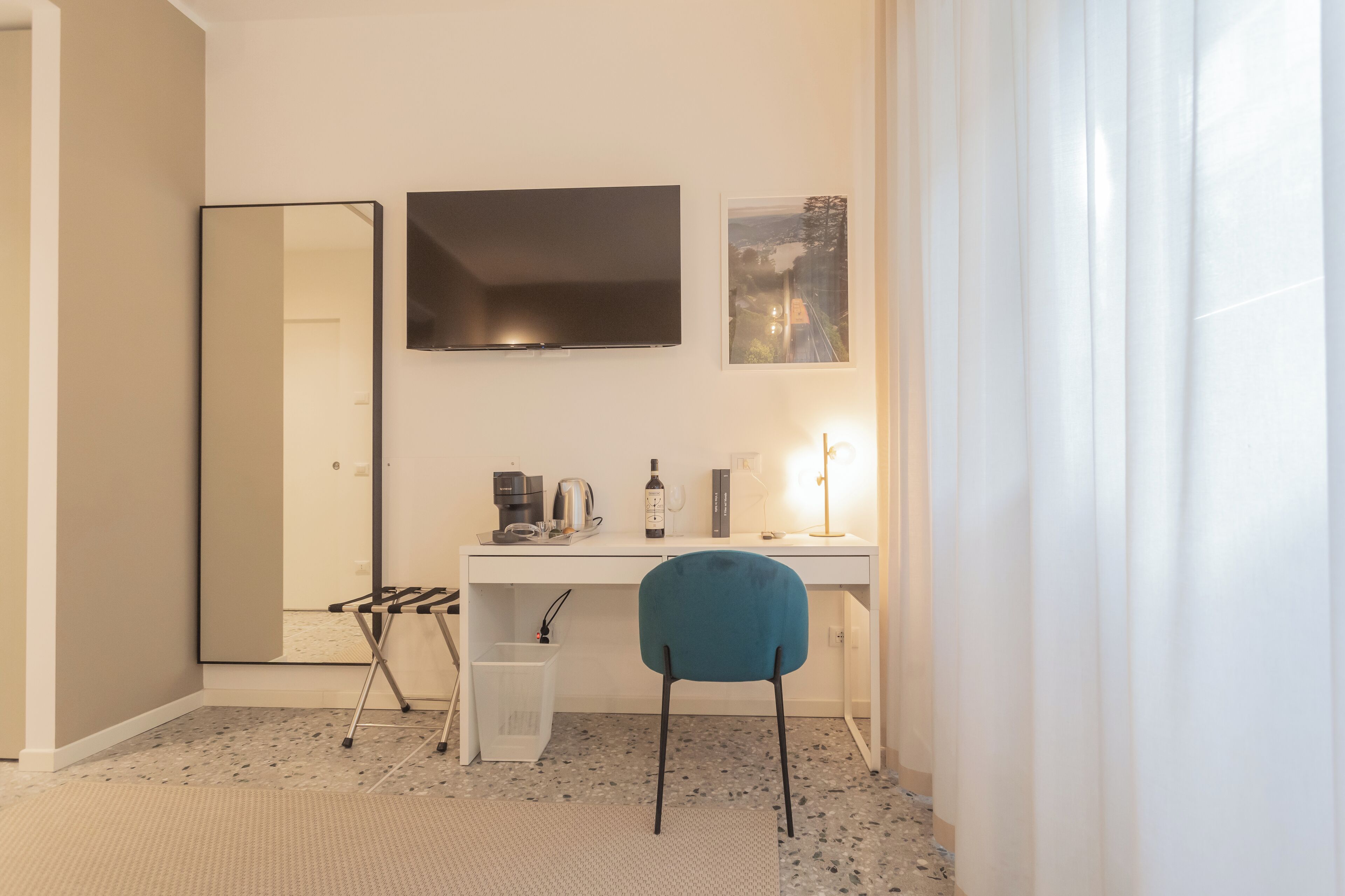 A modern hotel room with a white desk, a blue chair, a large mirror, and a framed picture on the wall.