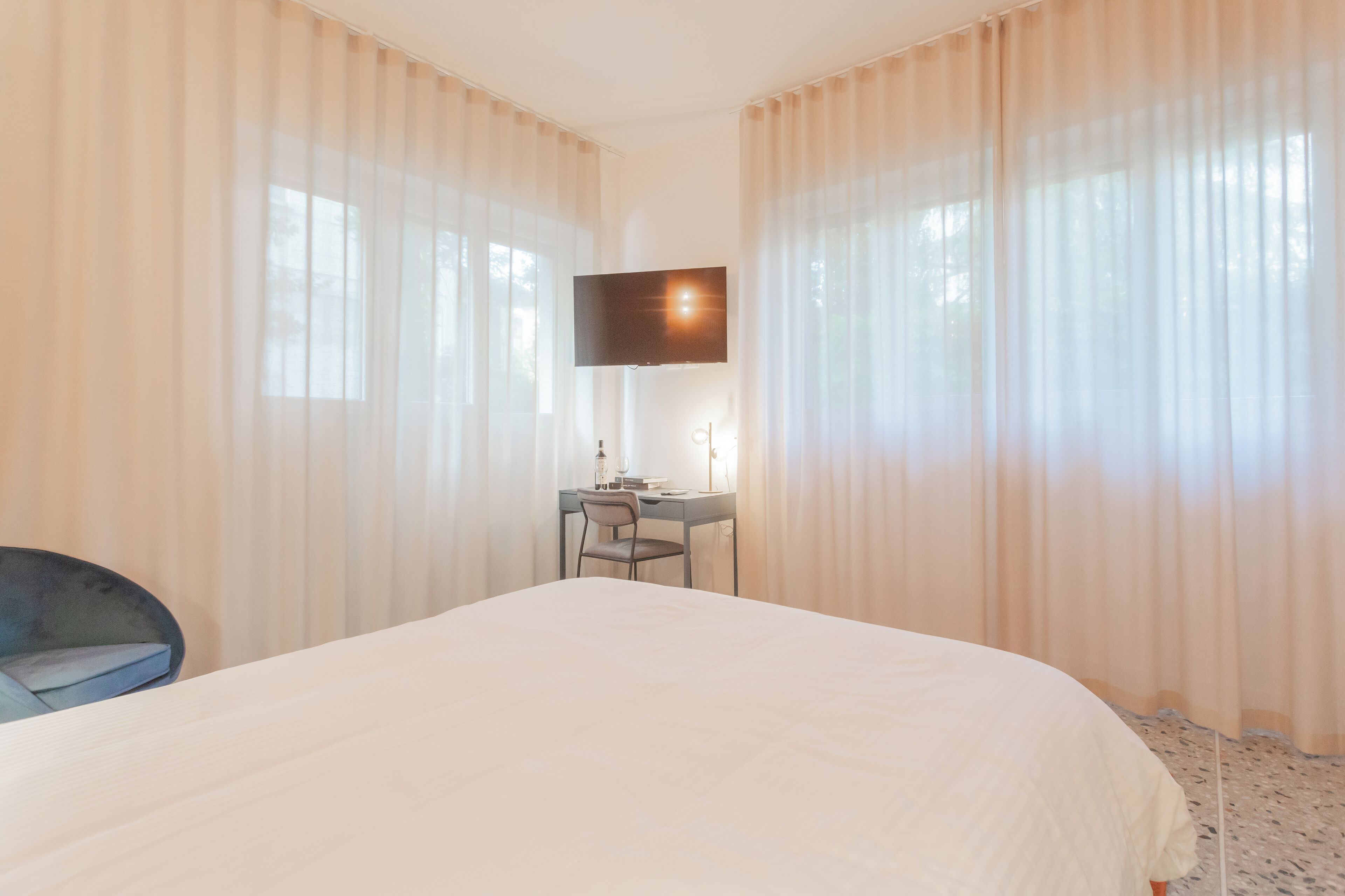 A hotel room with a large bed, a desk with a chair, a flat-screen TV mounted on the wall, and sheer curtains.