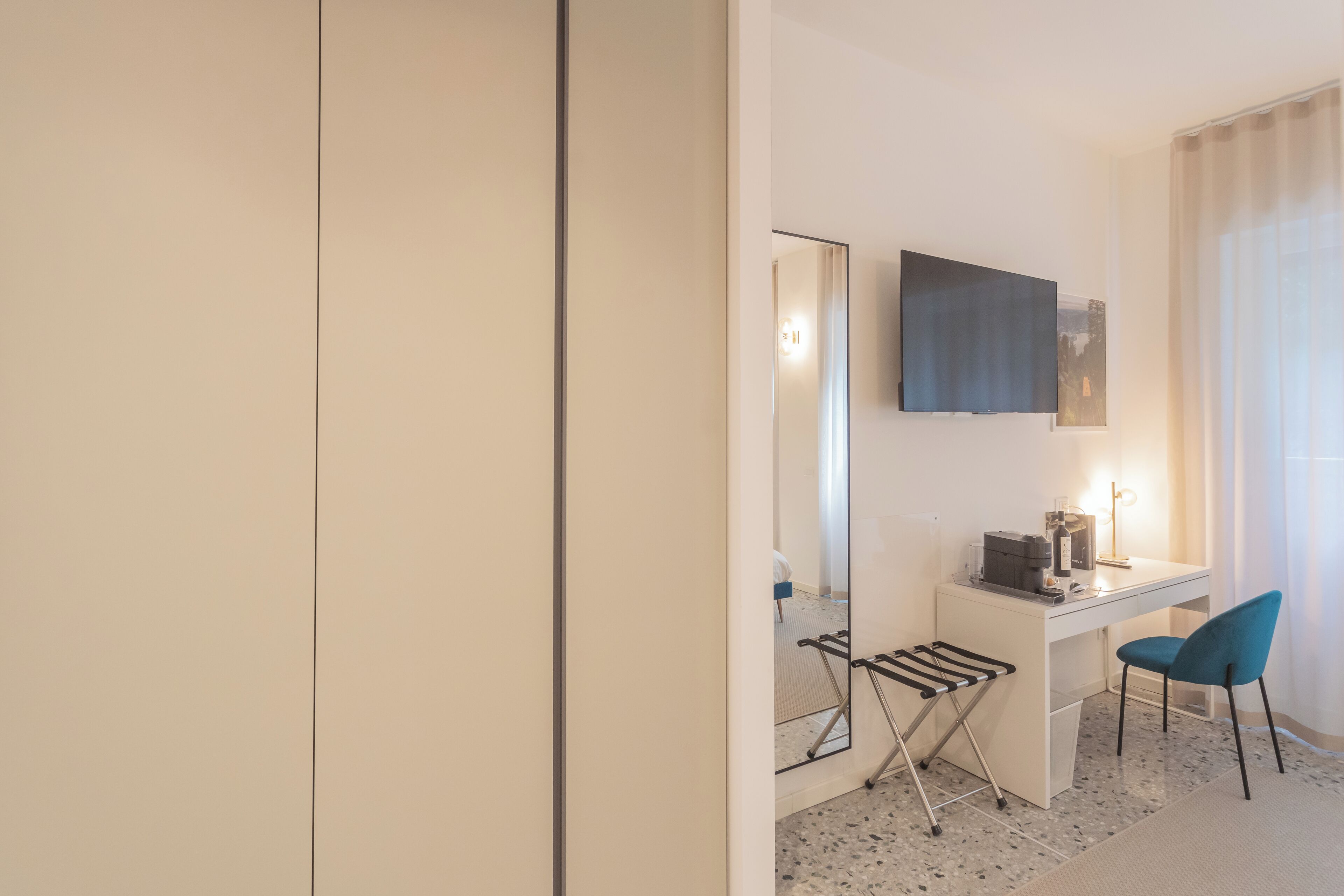 A modern hotel room with a white desk, a blue chair, a flat-screen TV, and a large mirror.