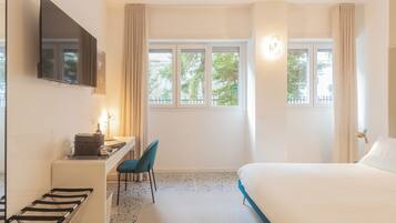 Habitación de hotel con cama, escritorio con silla, televisión montada en la pared y ventana con cortinas.