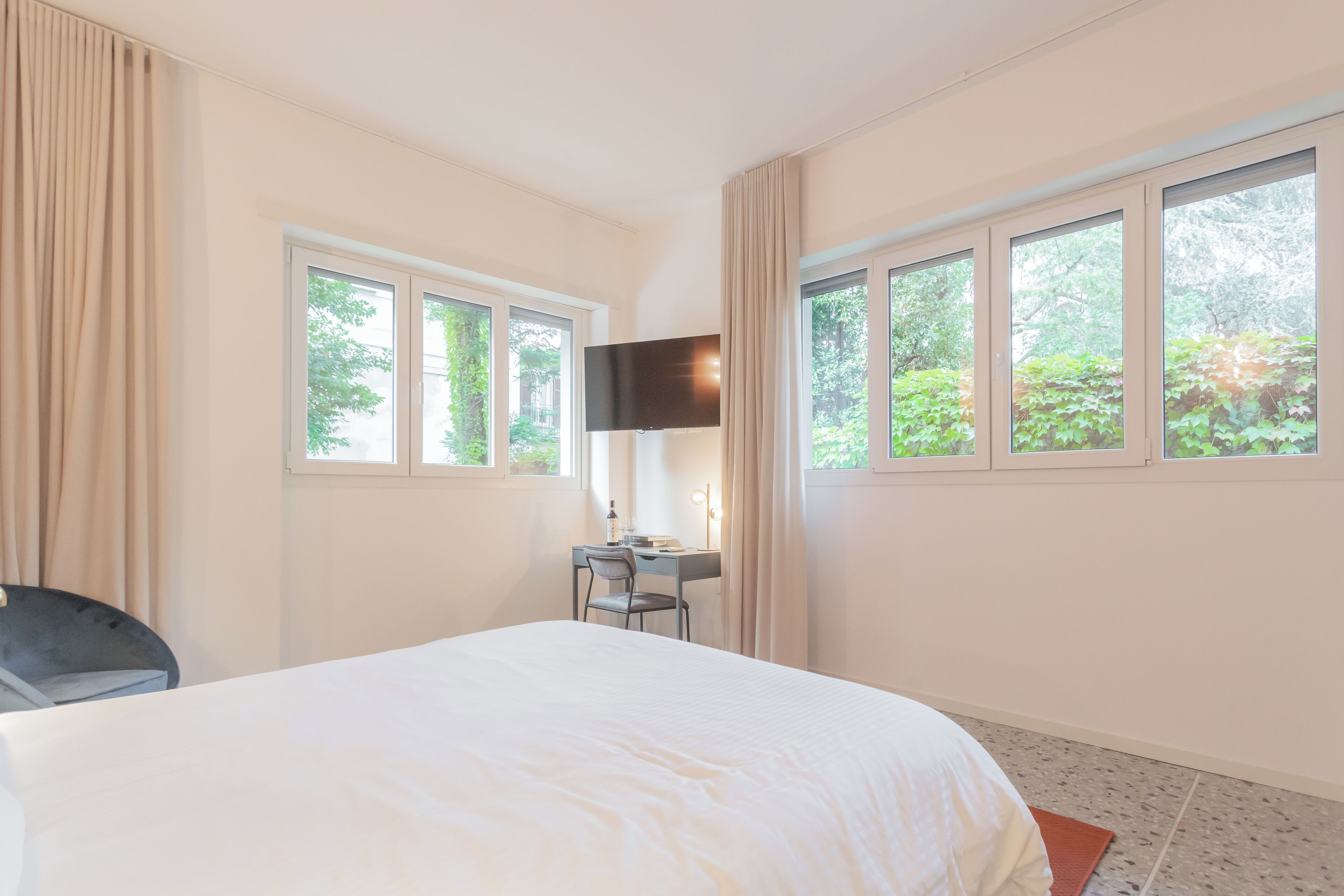 Hotelkamer met een bed, bureau, stoel, televisie en grote ramen met uitzicht op groen.