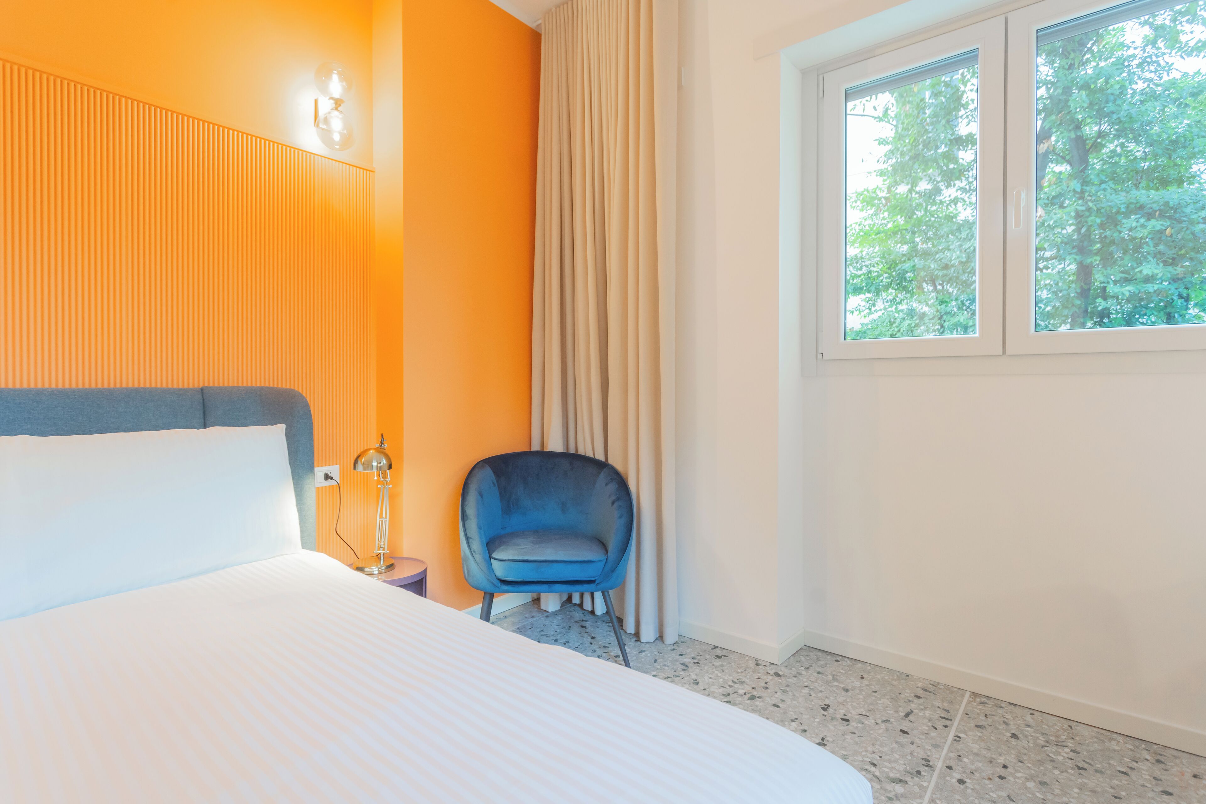 Une chambre d’hôtel avec un grand lit, une chaise bleue, une fenêtre donnant sur des arbres et un mur orné de lignes verticales.