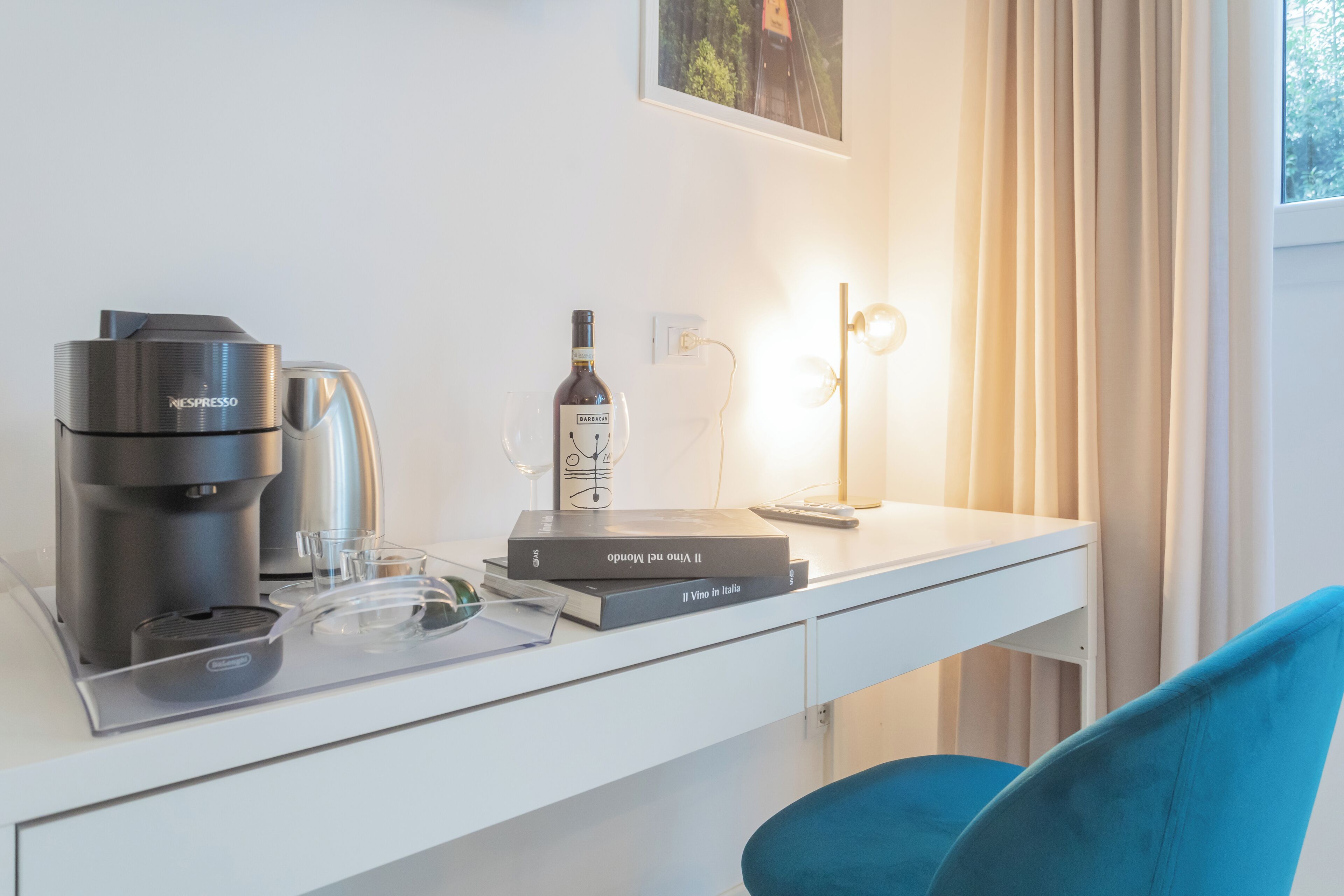 A modern hotel room with a white desk, a Nespresso coffee machine, a bottle of Barolo wine, and a framed picture on the wall.