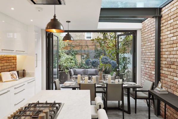 Une cuisine moderne dotée d’un îlot central, d’un coin repas avec chaises et tables, et offrant une vue sur un mur de briques et la verdure à l’extérieur.