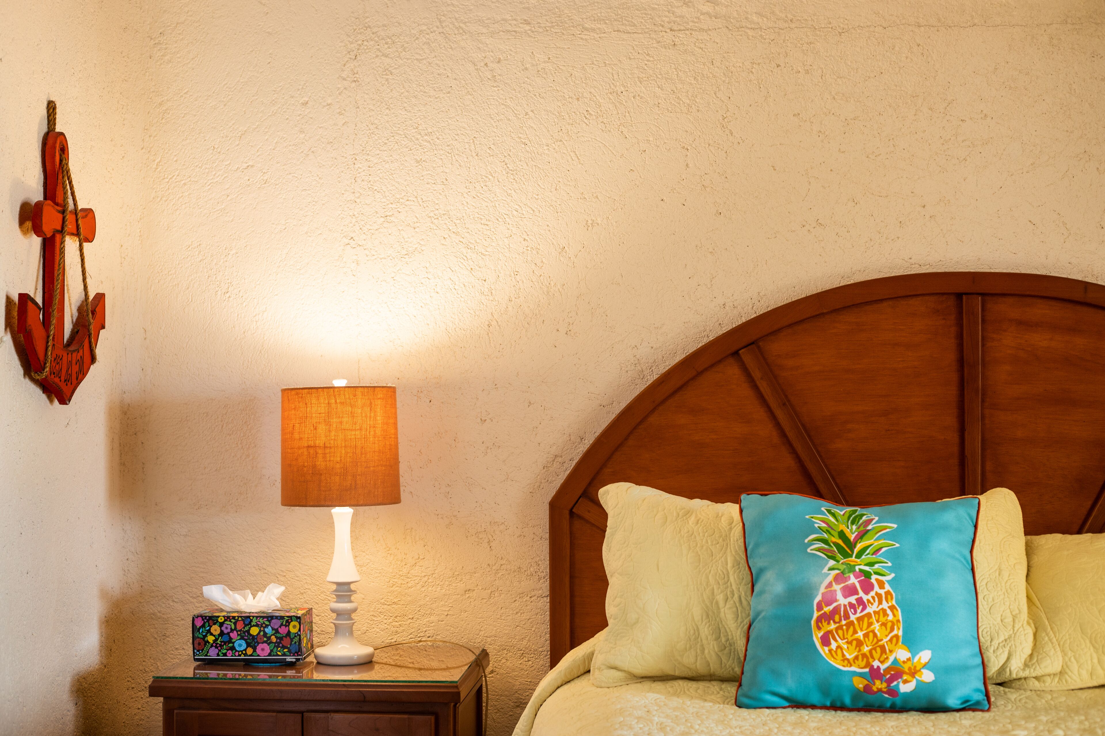 Een keurig opgemaakt bed met een houten hoofdbord, een bedlampje, een zakdoekendoos en een decoratief ananas kussen.