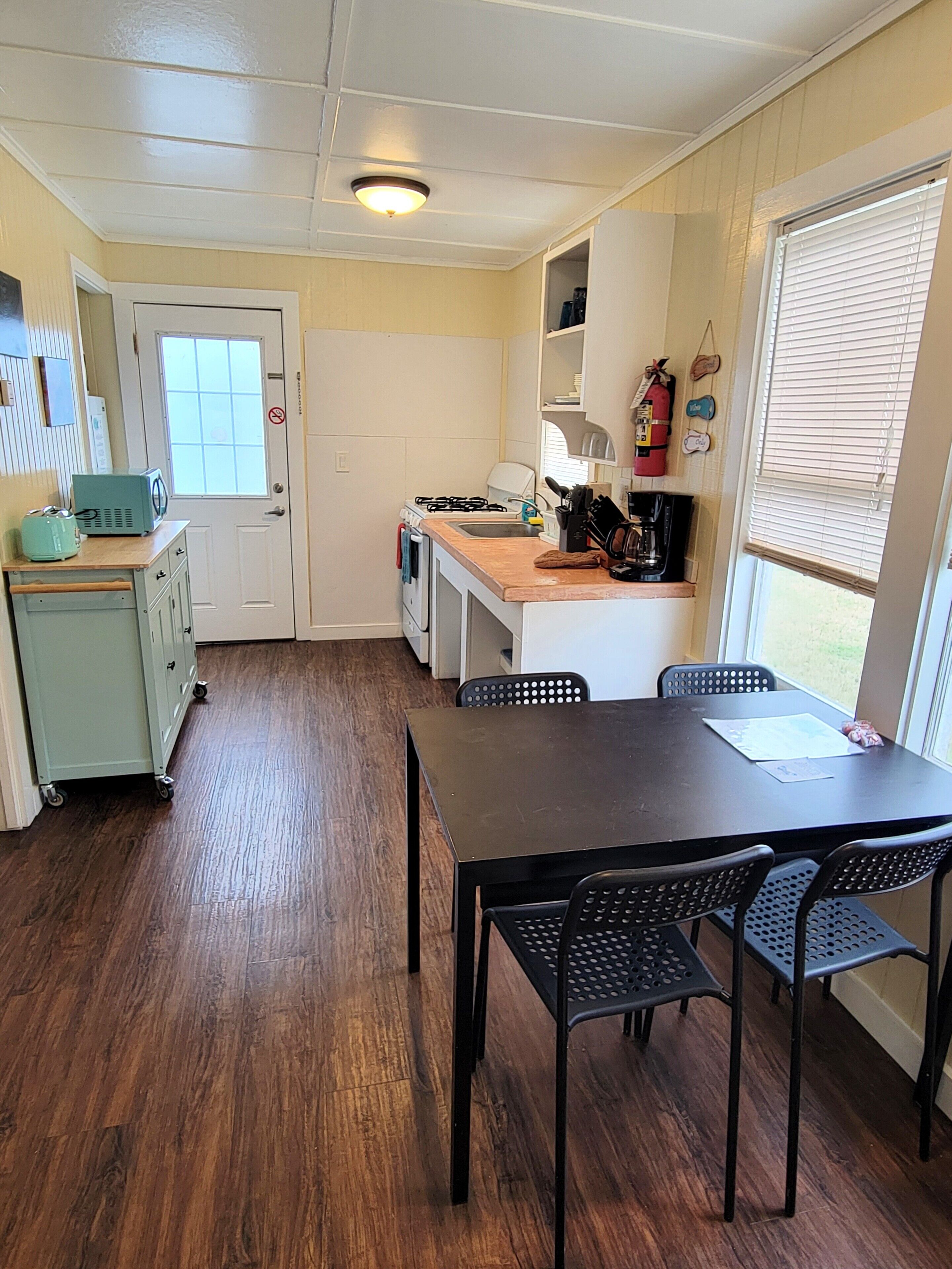 A compact kitchen with a dining area, wooden flooring, and a window with blinds.