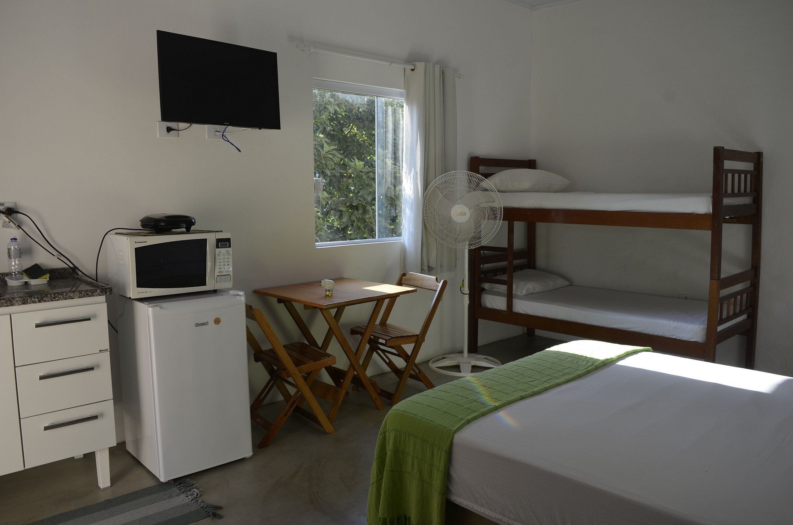 A compact room with a bunk bed, a small desk with a microwave, a chair, and a standing fan.