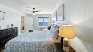 Une chambre à coucher comprenant un lit, un ventilateur de plafond, une table de chevet avec une lampe, une commode et une fenêtre donnant sur l’océan.