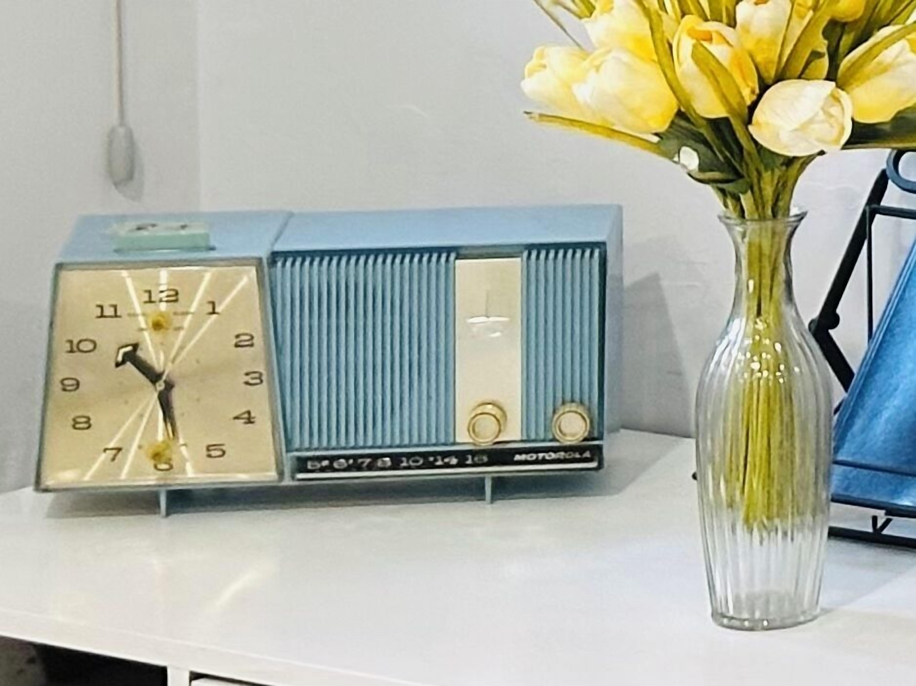 Una radio azul antigua con un reloj en la parte frontal, sobre una superficie blanca junto a un florero transparente con flores amarillas.