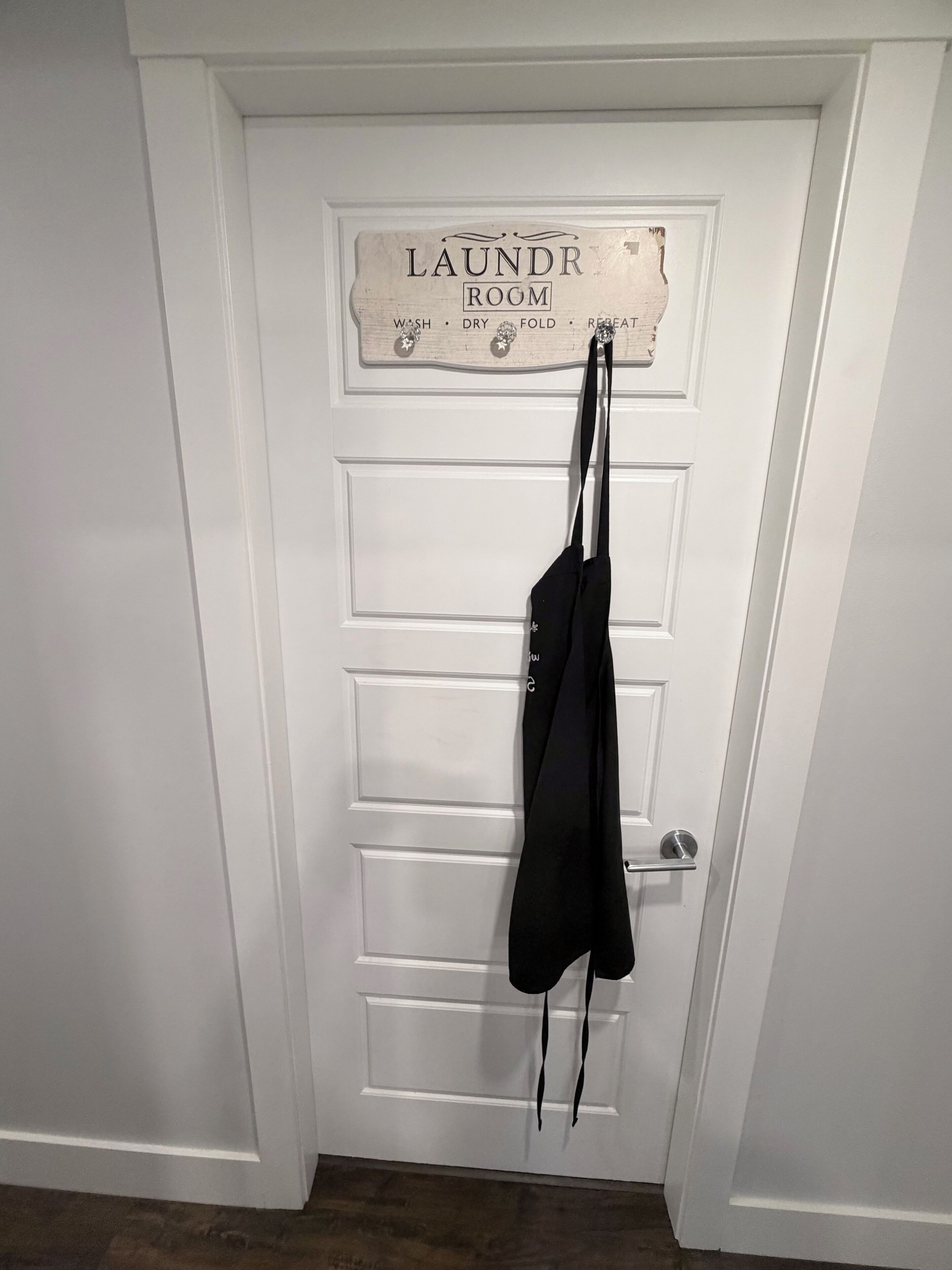 A white door with a sign above it that reads 'LAUNDRY ROOM' and lists steps 'WASH, DRY, FOLD, REPEAT.' A black garment is hanging on a hook to the right of the door.