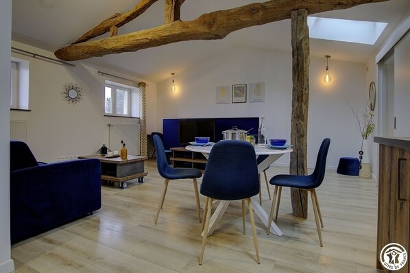 Una sala de estar moderna con comedor, que incluye una viga de techo de madera, un sofá, una mesa de comedor con sillas y una zona de cocina con televisor y armarios.