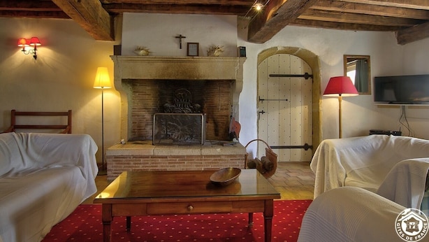 Ein gemütlicher Raum mit Kamin, zwei cremefarbenen Polstersesseln, einem Holztisch und einem roten Teppich mit Muster.