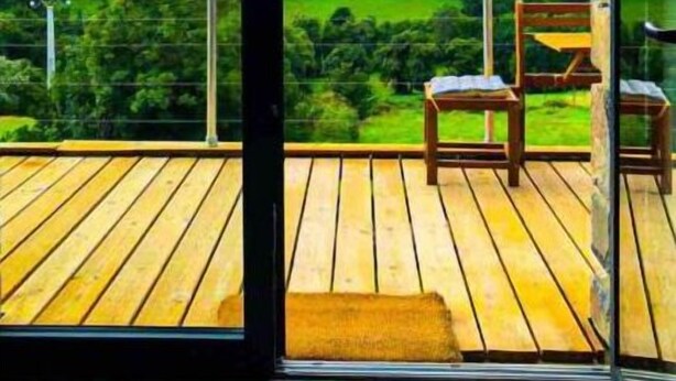 Un balcon doté d’un plancher en bois et de deux chaises, offrant une vue sur un paysage verdoyant avec des collines et un ciel nuageux.
