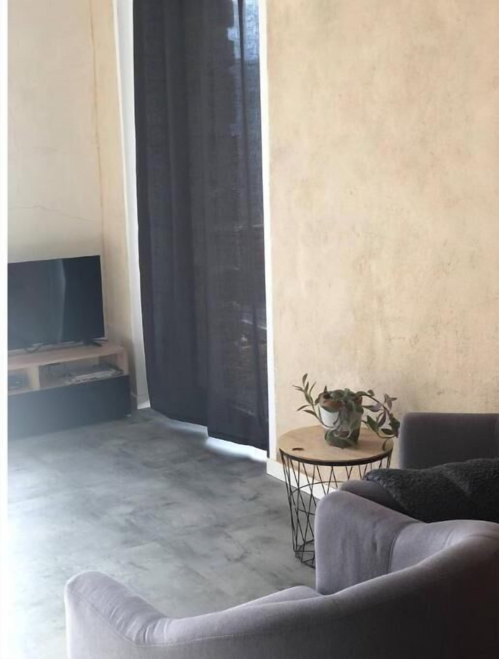 Una sala de estar con un sofá gris, una mesita redonda con una planta, un televisor sobre un mueble de madera y un ventanal grande con cortina oscura.