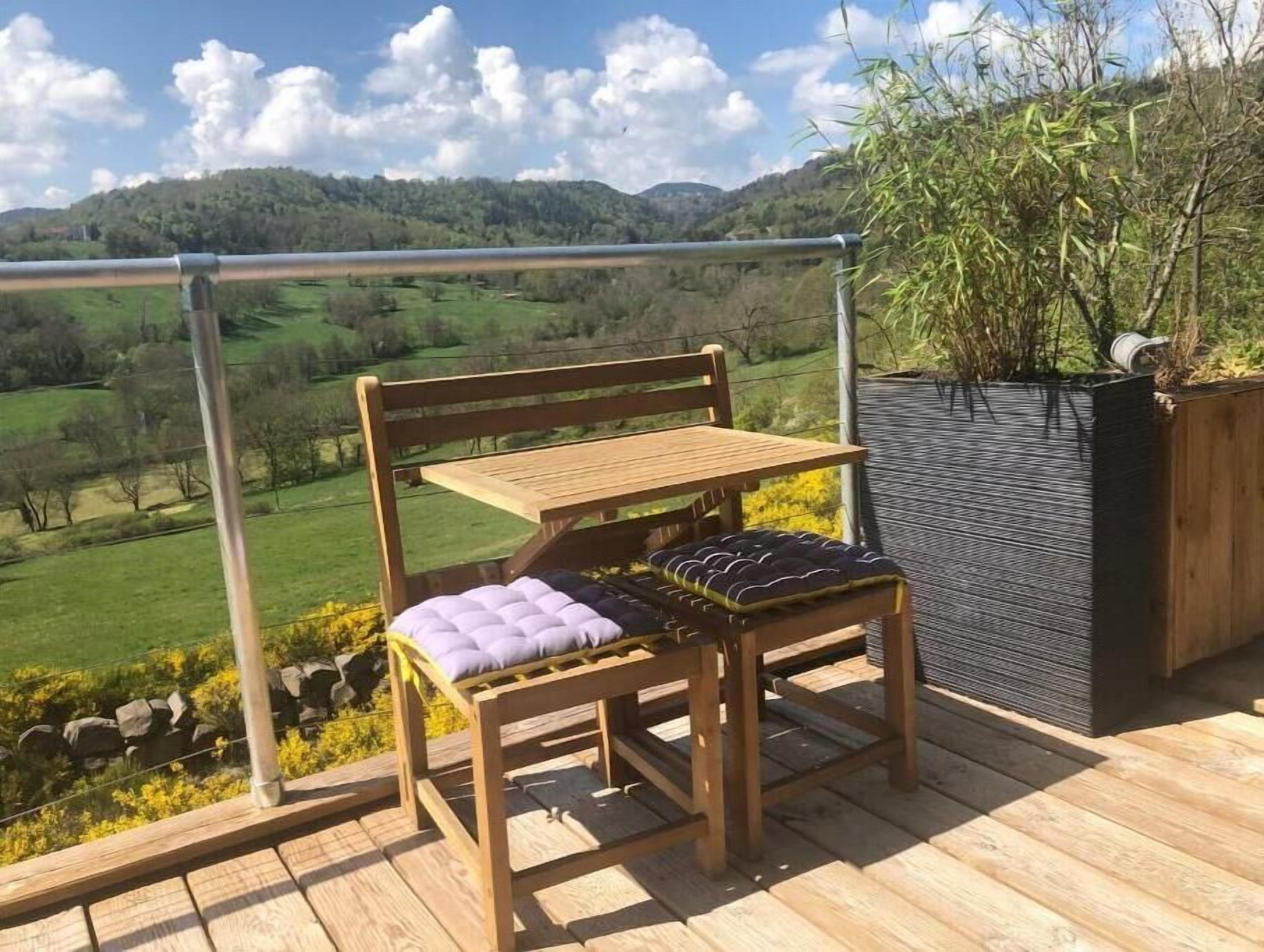 Une terrasse en bois avec un banc et une table, offrant une vue panoramique sur un paysage vallonné et un ciel dégagé.
