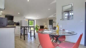 Une cuisine moderne avec des meubles blancs, un coin repas doté d’une table en verre et de chaises rouges, et un grand miroir au mur.