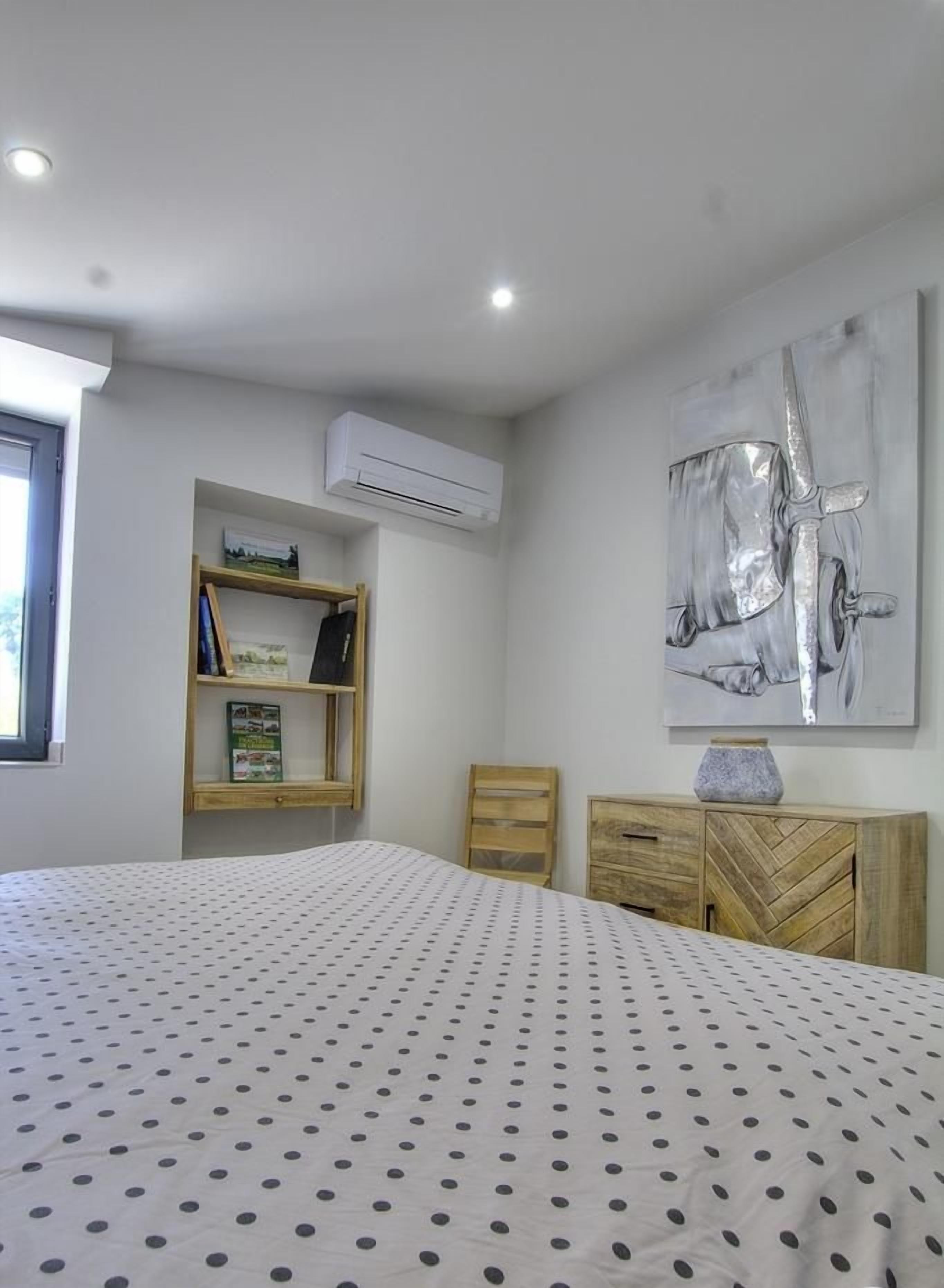 Een slaapkamer met een bed, een houten nachtkastje, een boekenkast en een wandairconditioner.