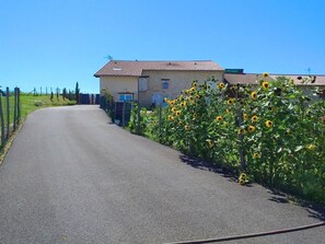 Une route de campagne, avec une maison sur la droite et des tournesols au premier plan.