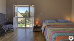 Een slaapkamer met een bed, een stoel, een nachtkastje met een lamp en een groot raam met uitzicht op de omgeving.