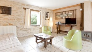 A room with a stone wall, a bed, a wooden table, a green chair, a television, and a window with curtains. - Le Tilleul - Courmangoux (COURMANGOUX)