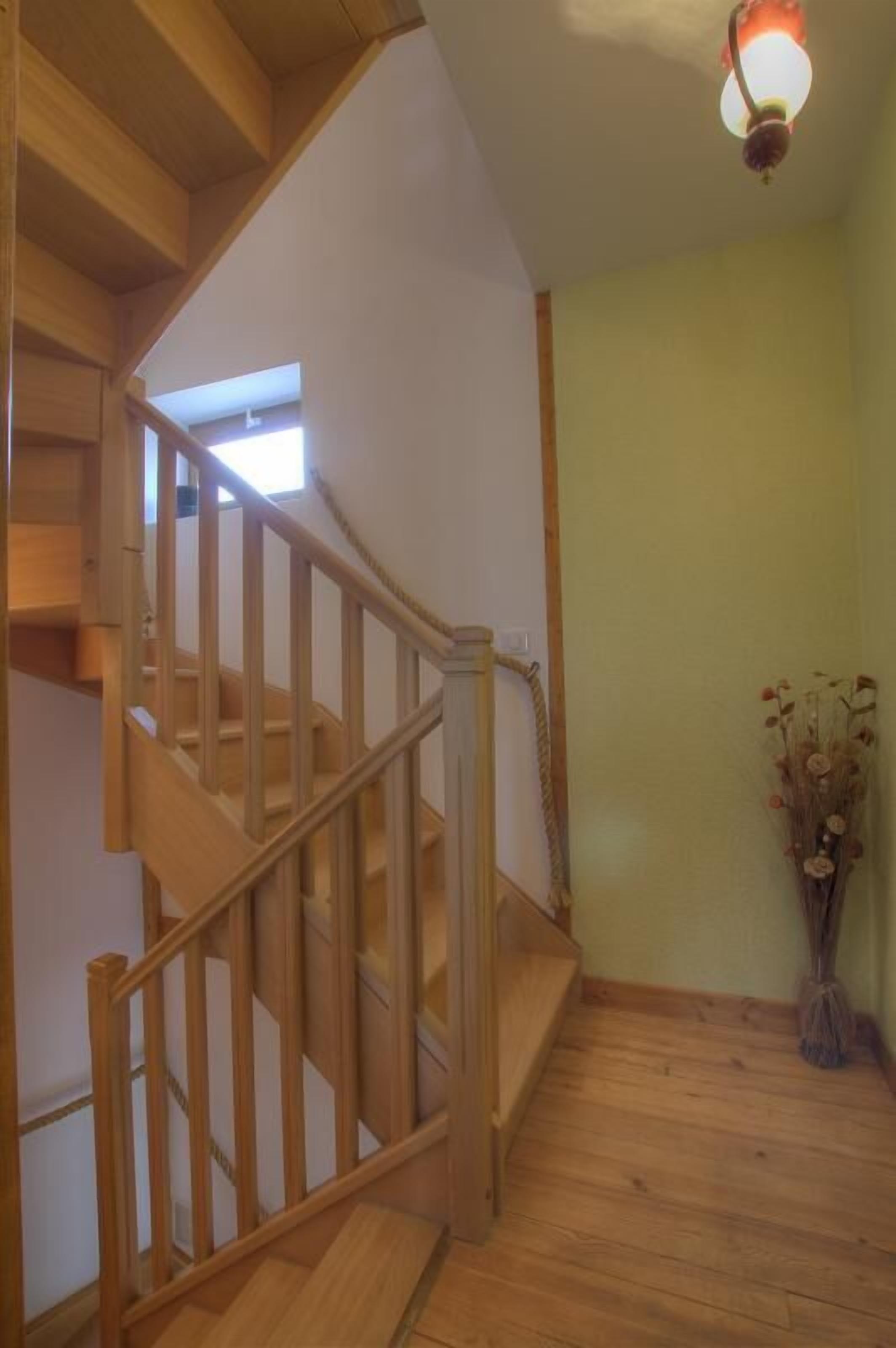 Un escalier en bois avec une rampe, un luminaire au-dessus et un vase rempli de fleurs séchées posé sur le sol.