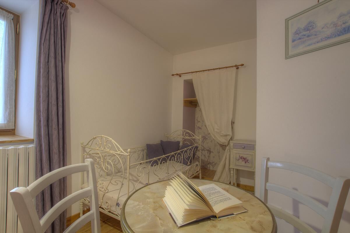 Una habitación pequeña con una cama de metal, una mesa redonda con un libro abierto y una ventana con cortinas.