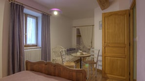 Une chambre à coucher avec un lit, une petite table ronde, deux chaises, une fenêtre avec des rideaux, une porte en bois et un luminaire au plafond.