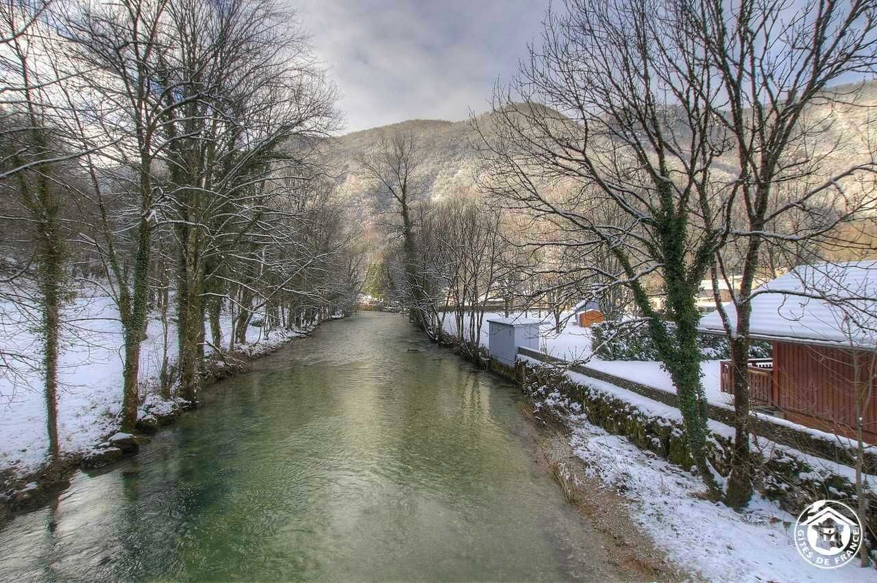 Ein schneebedeckter Fluss mit kahl stehenden Bäumen und einem Gebäude auf der rechten Seite.