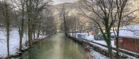 Ein schneebedeckter Fluss mit kahl stehenden Bäumen und einem Gebäude auf der rechten Seite.