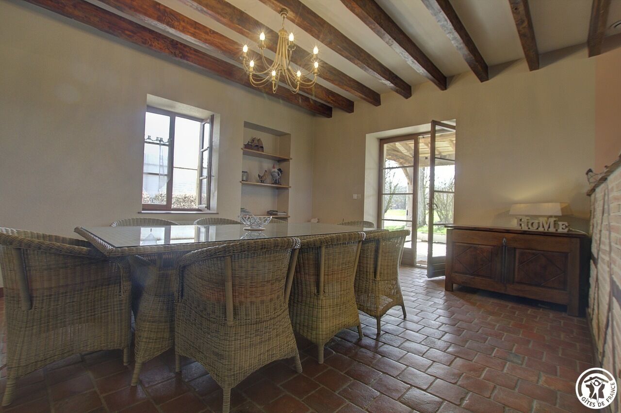 A dining area with a long table, wicker chairs, a chandelier, and a brick floor.