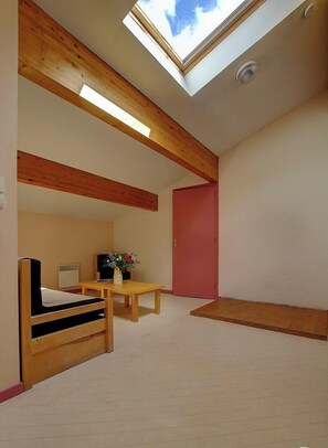 Una habitación con tragaluz, vigas de madera, una cama, una mesa con un jarrón y una puerta roja.