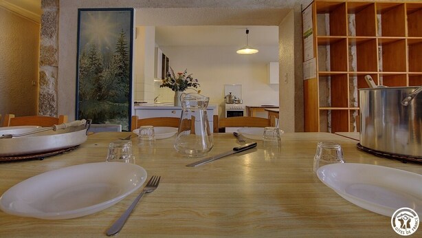 A dining area with a wooden table set for four, a painting on the wall, and a kitchen in the background.