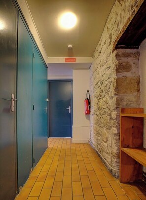 A hallway with a tiled floor, a stone wall, a blue door, and a fire extinguisher on the wall. - Gite Communal - Plateau D'hauteville (PLATEAU D'HAUTEVILLE)