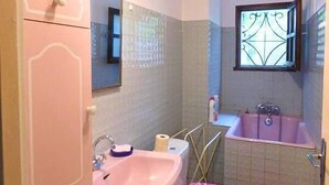 Une salle de bain avec un lavabo rose, des toilettes avec un abattant rose, une baignoire et une fenêtre ornée d’un motif décoratif.