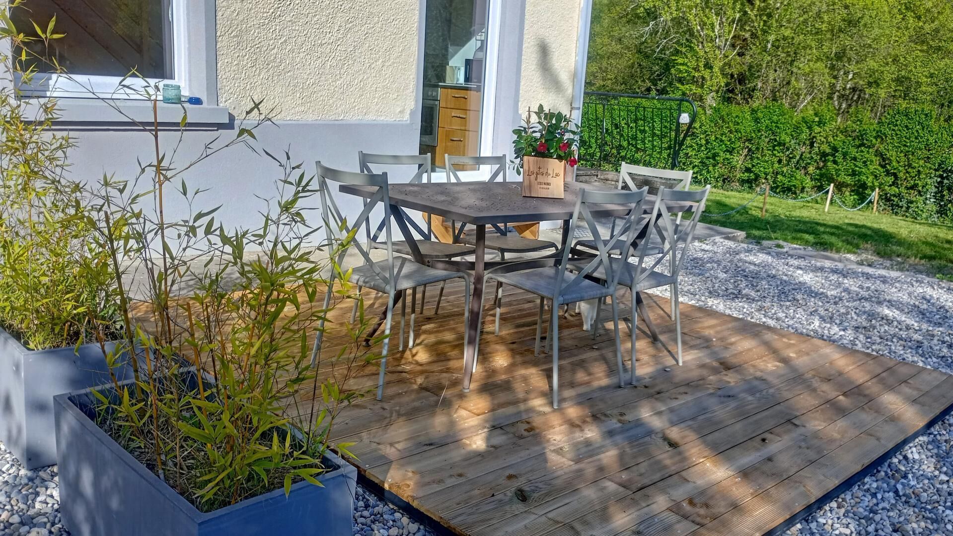 Eine Holzterrasse im Freien mit Tisch und Stühlen, umgeben von Blumentöpfen und einer Hecke.