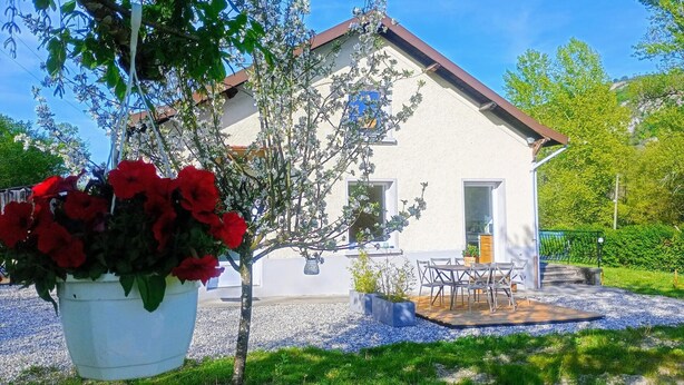 Ein Haus mit weißer Fassade, einem braunen Dach und einer Zuwegung mit Kies. Vor dem Haus befindet sich eine Holzterrasse mit Tisch und Stühlen, im Vordergrund ein Blumentopf mit roten Blüten.